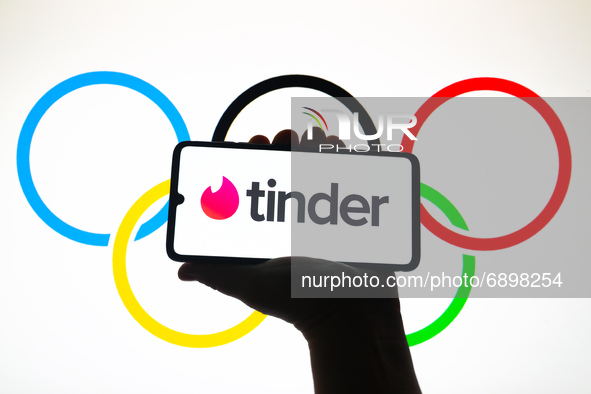 Tinder logo is displayed on a mobile phone screen photographed with Olympic Rings symbol background for illustration photo. Krakow, Poland o...