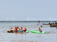 Holidaymakers enjoy sunny summer day at the beach in Whitstable, south-eastern coastal town of England with Kentish Flats windmills power pl...