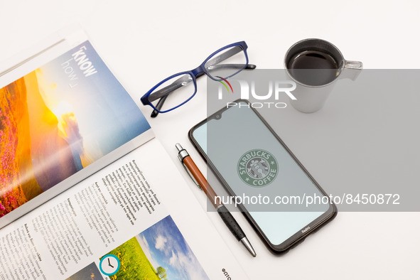 In this photo illustration a Starbucks Coffee logo seen displayed on a smartphone screen on a desk next to a cafe, a pen, glasses and a maga...