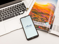 In this photo illustration an Alaska airlines logo seen displayed on a smartphone screen on a desk next to a Macbook and atravel magazine in...
