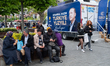 People sitting and passing by on the bench next to the poster showing the portrait of Rece...