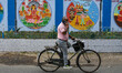A man is cycling past a wall decorated with various depictions of a Bengali festival, as s...