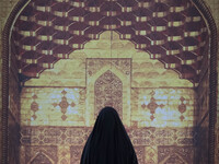 A veiled Iranian woman is standing in front of a massive banner featuring an image of the Imam Hussein holy shrine in Karbala, inside the Im...