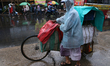 A street food vendor covered with a plastic sheet with his cycle-loaded goods crosses a bu...