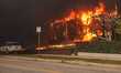 Flames engulf a residential structure, with nearby vehicles parked along a smoke-filled st...