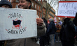 A protester holds a cardboard sign reading 'Judo-leftist'. The placard behind reads 'Anti...