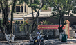 A woman wearing a helmet and face mask sits on a motorbike parked in front of a government...