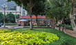 A small garden with trees, flowerbeds, and a path leads to a row of souvenir shops in cent...