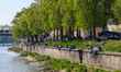 People sit and relax on the stone embankment along the Danube River beneath a row of leafy...