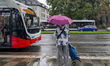 A person with an umbrella and a shopping trolley stands during rain at a bus stop while a...
