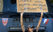 A young man stands in front of a water cannon with a cardboard sign reading 'Cut yours, th...