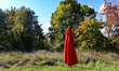 A closed red parasol stands on a grassy field surrounded by trees with early autumn foliag...