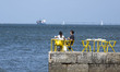 Two people sit near a port area on the Tejo River, while in the background a cargo ship an...