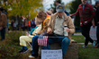 An older man holding an American flag and a water bottle sits beside a sign reading ''The...
