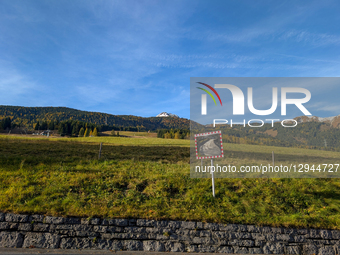 A traffic safety mirror, framed in red and white checkers, reflects a winding road, set against a backdrop of green fields, autumnal forest,... by Michael Nguyen/NurPhoto