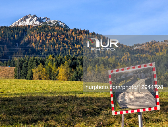 A traffic safety mirror, framed in red and white checkers, reflects a winding road, set against a backdrop of green fields, autumnal forest,... by Michael Nguyen/NurPhoto