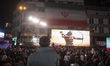 Iranian people sit under a country flag and a display depicting a symbolic image of the Ir...