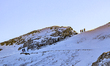 Two small groups of hikers ascend a wide, steep, snow-covered mountain slope on a clear da...