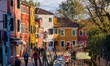 People walk along the waterway past moored boats and colorful residential buildings with l...