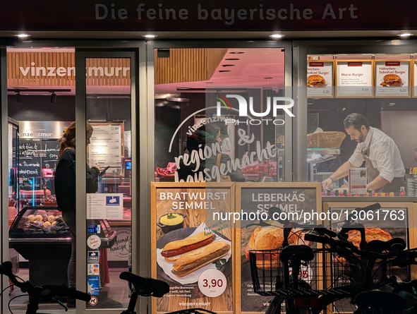 The illuminated storefront of Vinzenzmurr displays meat, sausage products, and sandwich advertisements. A clerk serves a customer inside, wh... by Michael Nguyen/NurPhoto