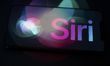 The Siri logo appears on the screen of a smartphone placed on a surface reflecting the new...