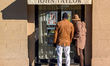 A couple stands in front of a luxury real estate agency John Taylor window displaying prop...