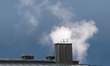 Smoke rises from a residential chimney above an apartment building under dark storm clouds...