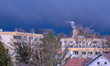Smoke rises from a residential chimney above an apartment building under dark storm clouds...