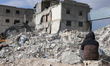 An elderly woman sits on the ruins of her destroyed home, waiting for her family to be pul...