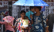 A woman carries her baby and blows air with one hand while walking on a hot summer day in...
