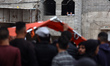 Palestinians look through a shattered window as mourners carry the body of a Palestinian d...