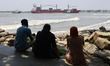 Beachgoers sit on a seaside promenade as a cargo ship passes through the Arabian Sea in th...