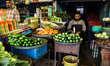 A shopkeeper sells vegetables while another sells cooking oil, juices, and other grocery i...