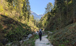 Two hikers walk with trekking poles along a forest trail beside a stream in Berchtesgaden,...