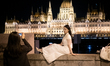 A tourist sits on a wall by the Danube River and poses for a photo with the illuminated Hu...