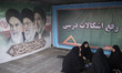 A group of veiled Iranian women sit together beside a banner depicting portraits of Iran's...