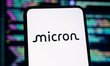 The Micron Technology logo is displayed on a smartphone screen with lines of computer code...