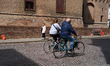 Two unidentified cyclists ride along the cobblestone street beside the medieval brick wall...