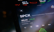 The SpaceX stock ticker is displayed on a smartphone screen placed on a reflective surface...