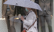 A woman with her face mask on holds an umbrella as she walks past a shop window with manne...