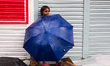 A little girl takes shelter from rain standing with her umbrella, outside a closed shop at...