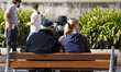 An old man with face mask and a hat talks with people on a bench during a sunny and warm V...