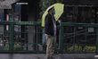 A man carries an umbrella while waiting for a bus in front of the area where a column of t...