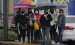 A group of women carry umbrellas while waiting for a bus in front of the area where a colu...