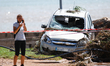A woman speaking on the phone in front of a car damaged by floods on street filled with mu...