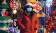Woman earing a santa hat and face mask stands by Christmas decorations outside a house dur...