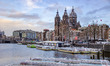 The canals and parked boats in Amstedam with the Basilica of Saint Nichola in the backroun...