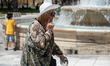 A woman wearing a hat for protection and holding a bottle of water walks past a fountain a...
